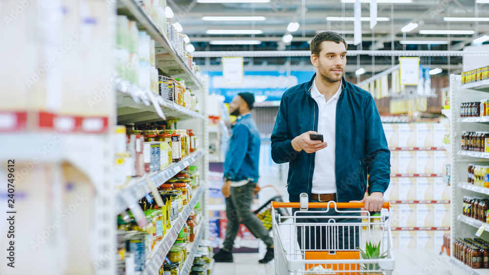 At the Supermarket: Handsome Man Uses Smartphone and Browses Through the Canned Goods Shelf. He's Standing with Shopping Cart in Canned Goods Section.