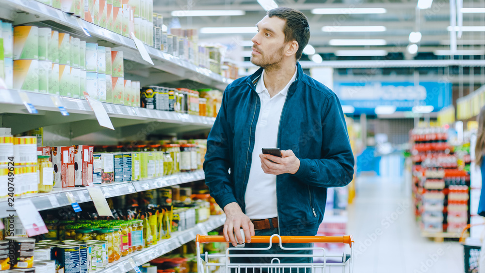 At the Supermarket: Handsome Man Browses Through Shelf with Canned Goods, Places Tin Can into His Shopping Cart and Proceed with His Shopping List Walking Through Different Sections.