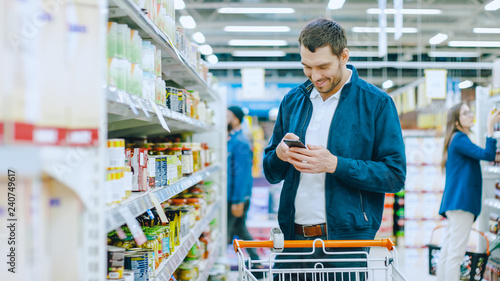 At the Supermarket: Handsome Man Uses Smartphone and Stands in Canned Goods Section. He's Looking for a Best Healthy Product.