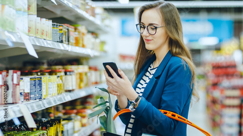 At the Supermarket: Beautiful Young Woman Uses Smartphone While Browsing through the Canned Goods Section of the Store. She Checks Her Shopping List and Holds Shopping Basket. photo