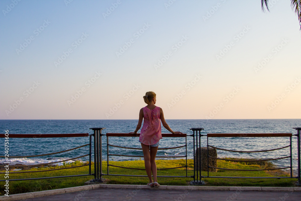 Fototapeta premium Young slender girl stands on the promenade by the sea, hot summer weather, sunset. City Side, Turkey.