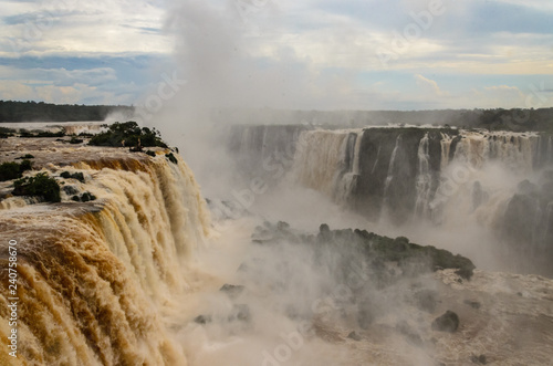 Iguazu falls on the brazilian side with some green bushes, water mist and a cloudy sky