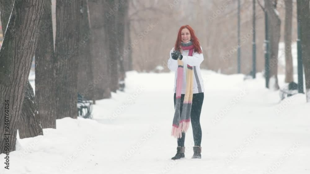 Winter park. A cheerful woman with bright red hair standing on the sidewalk, making a snowball and throwing it forward. Slow motion Winter park. A cheerful woman with bright red hair standing on the sidewalk, making a snowball and throwing it forward. Slow motion