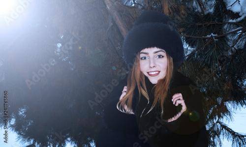 Portrait of a beautiful young girl in a black hat on the background of dark pine branches in winter the sun shines. © YAROSLOVEPHOTOVIDEO