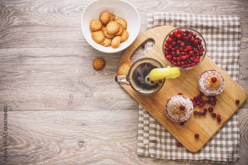 On a light wooden tabletop on a linen napkin napkin, there is a cutting board with two muffins, a broken chocolate bar and bright red berries in a small tree, next to a bowl with cookies. On a light wooden tabletop on a linen napkin napkin, there is a cutting board with two muffins, a broken chocolate bar and bright red berries in a small tree, next to a bowl with cookies.