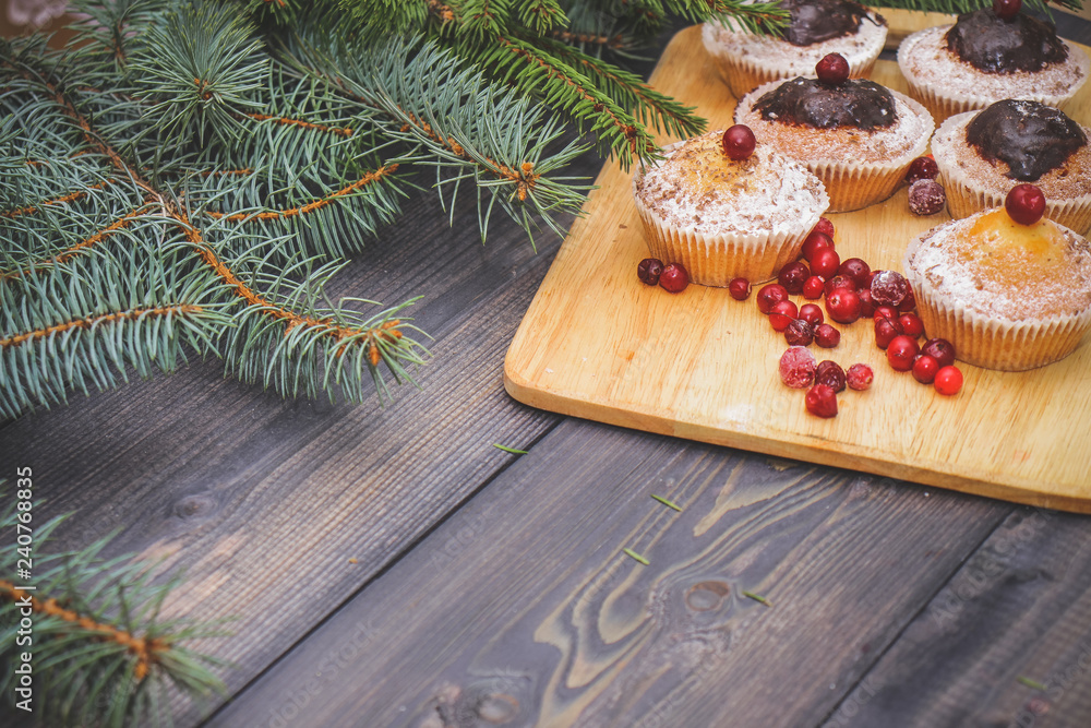 On a gray wooden tabletop of a blue spruce branch, on a light wooden cutting board, muffins, decorated with powdered sugar and chocolate, berries of red cranberries. On a gray wooden tabletop of a blue spruce branch, on a light wooden cutting board, muffins, decorated with powdered sugar and chocolate, berries of red cranberries.