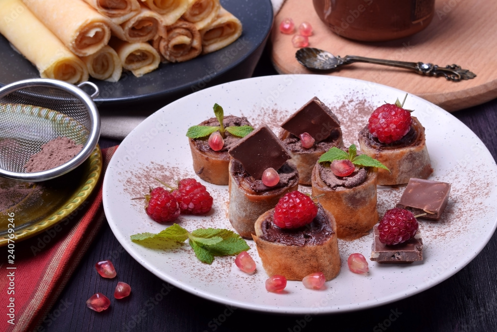 Rolled crepes with chocolate paste topped with raspberries, pomegranate, mint and chocolate pieces on a white plate