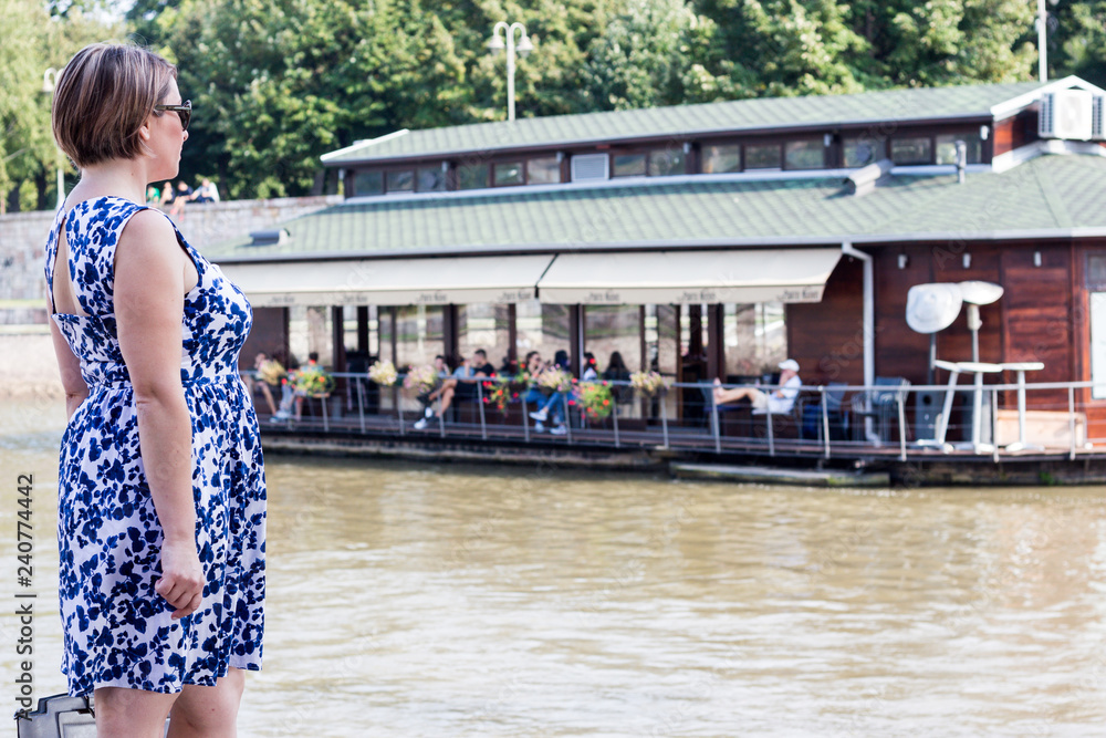 Beautiful Brunette Girl in a Blue Dress Posing Next to the River and Looking at the River Cafe Boat