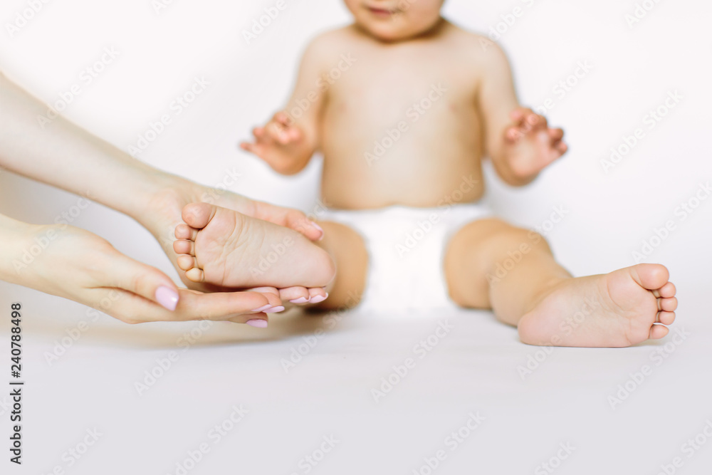 Mother massaging her child's foot, cute little baby girl in white diaper is sitting on the white background. Baby health and hygiene concept Mother massaging her child's foot, cute little baby girl in white diaper is sitting on the white background. Baby health and hygiene concept