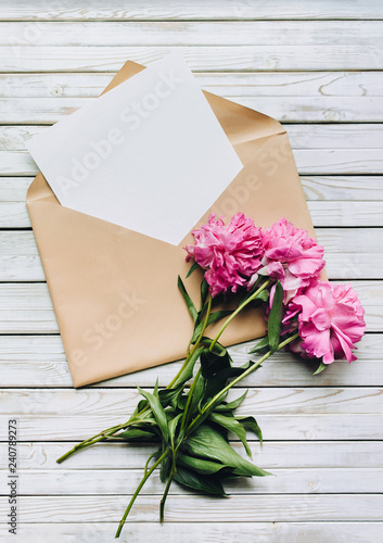 Faded purple peonies and an open kraft envelope containing a white sheet of paper. Valentine's Day concept. Copy space, top view.