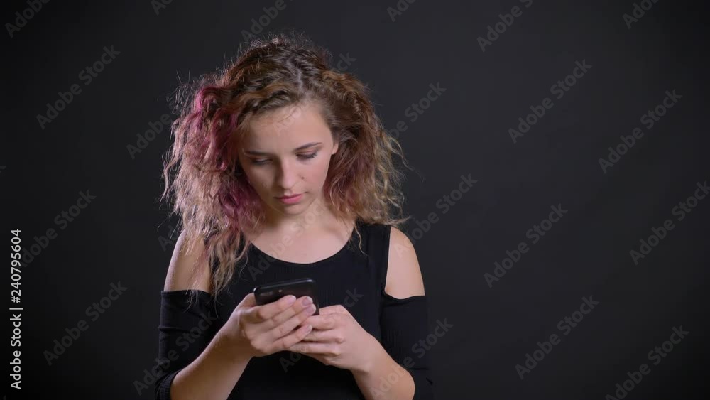 Portrait of young caucasian girl with pink hair attentively watching into her smartphone on black background. Portrait of young caucasian girl with pink hair attentively watching into her smartphone on black background.