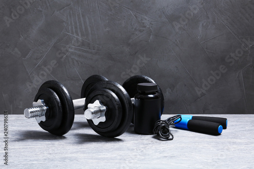 Fototapeta Naklejka Na Ścianę i Meble -  Pair of adjustable dumbbells, jump rope and jar with protein on floor near gray wall. Space for text