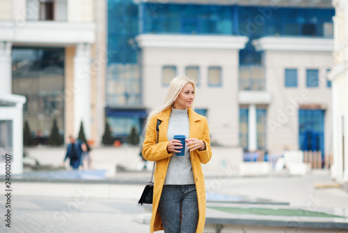 Coffee on the go. Beautiful young blond woman in bright yellow coat holding coffee cup and smiling while walking along the street 