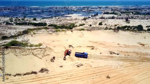 aerial view sand quarry with truck by excavator and artificial ponds against ocean under bright blue sky