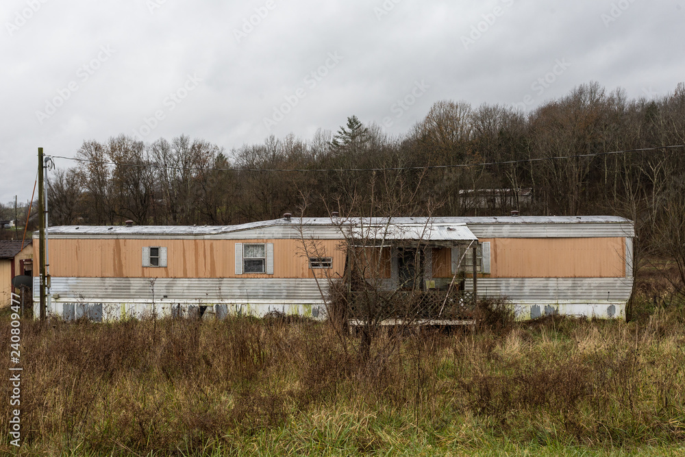 Abandoned mobile home with orange and white trim surrounded by overgrown grass