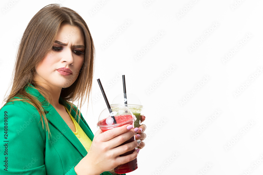 Healthy Food Eating. Choosy Thinking Woman With Both Green and Red Detox Vegetable Smoothie. Posing in Green Jacket Over White.Unpleased Facial Expression. Healthy Food Eating. Choosy Thinking Woman With Both Green and Red Detox Vegetable Smoothie. Posing in Green Jacket Over White.Unpleased Facial Expression.