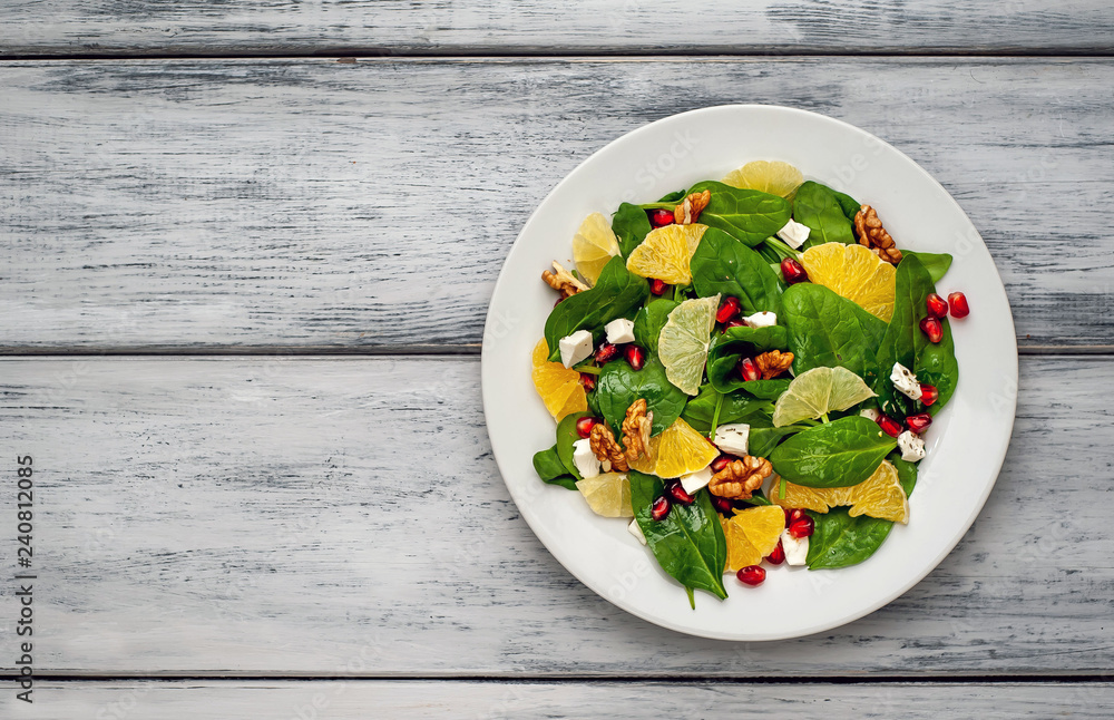 Spinach salad, orange, lemon, pomegranate, feto cheese, walnut on a white plate on a wooden table