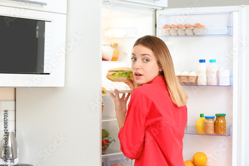 Emotional young woman taking sandwich from refrigerator in kitchen. Failed diet