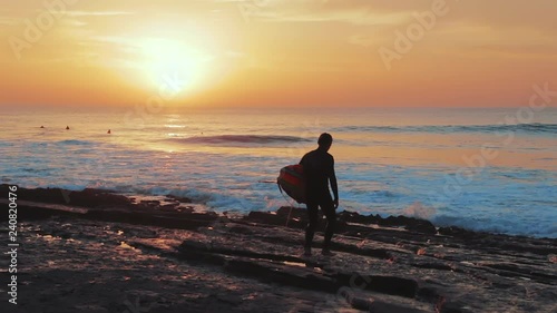 A surfer walks towards the ocean with surf in his arms with the sun setting in the background. Surf break and lineup in the background.