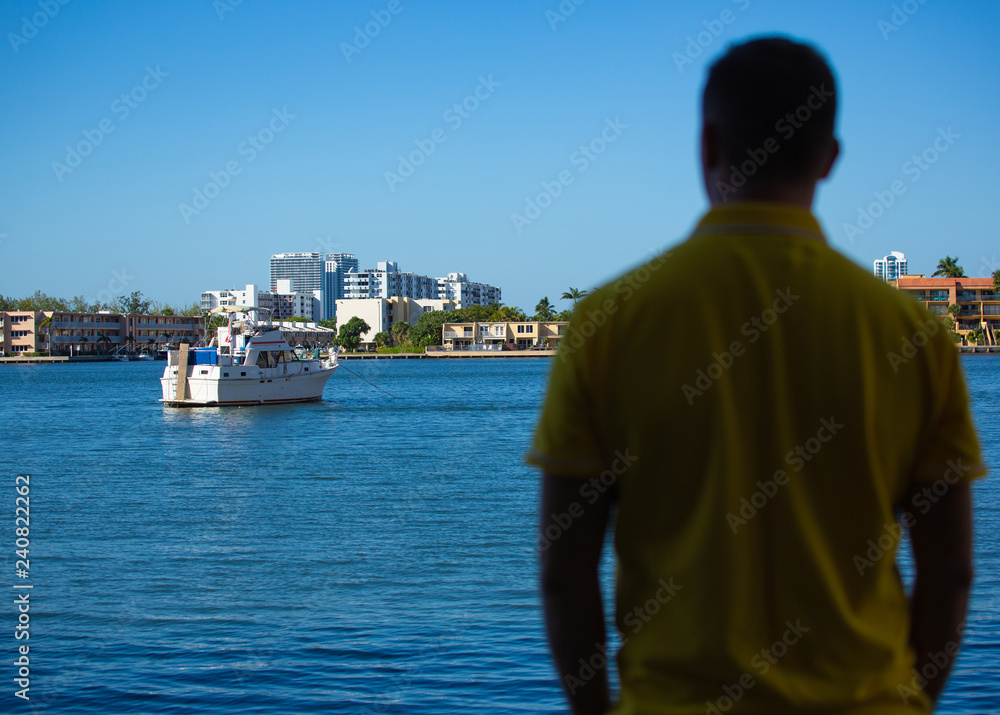 A man in a yellow polo  standing with his back to the camera and looks at the water and modern boat