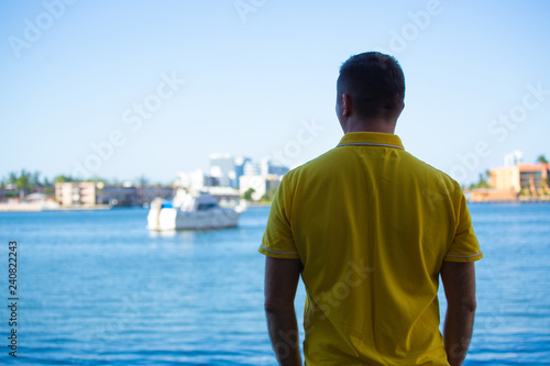 A man in a yellow polo  standing with his back to the camera and looks at the water and modern boat