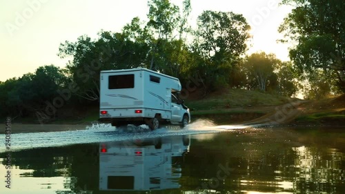 A caravan drives into view and stops before a hill leading up the sunset and heavily forested bushland.