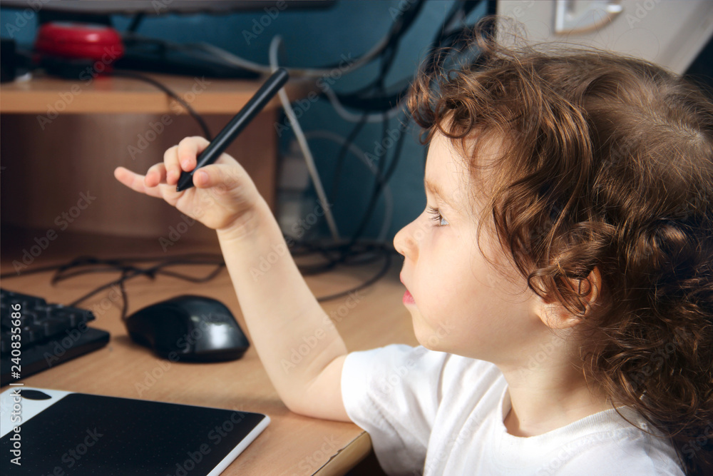 Little 2 3 year old baby girl in white clothers draws at the home computer in graphics drawing tablet. The child is holding a pen and laughs. On the head are huge headphones