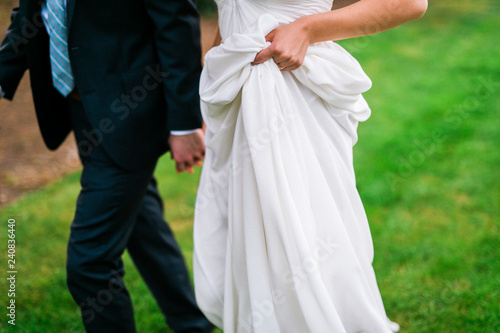 bride holding dress while walking with groom in grass