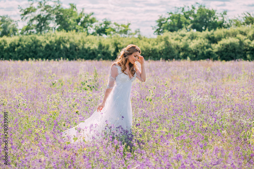 lovely sweet girl with closed eyes, dressed in an elegant long white dress with transparent sleeves, touches a neat hairstyle of lond hair, walks across the field of lilac purple flowers
