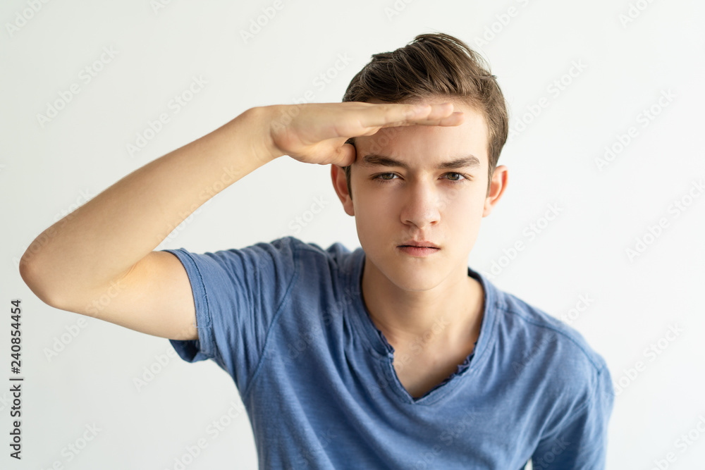 Concentrated young man looking into distance. Serious frowning guy in blue tshirt holding hand near forehead and looking at camera. Choosing goals concept Concentrated young man looking into distance. Serious frowning guy in blue tshirt holding hand near forehead and looking at camera. Choosing goals concept