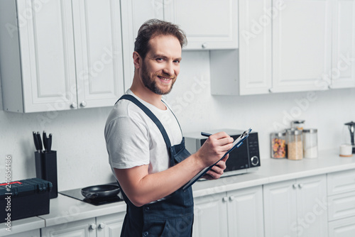 happy adult bearded repairman making notes in clipboard in kitchen