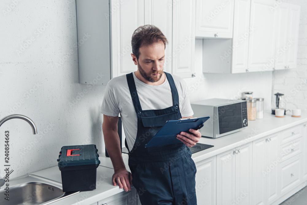 adult bearded repairman looking at clipboard in kitchen at home