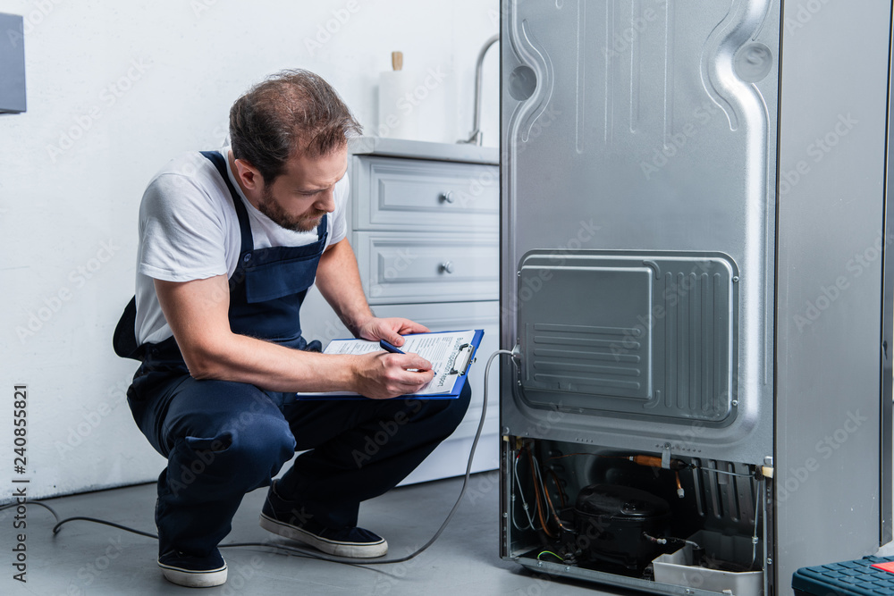 adult craftsman in working overall writing in clipboard near broken refrigerator in kitchen