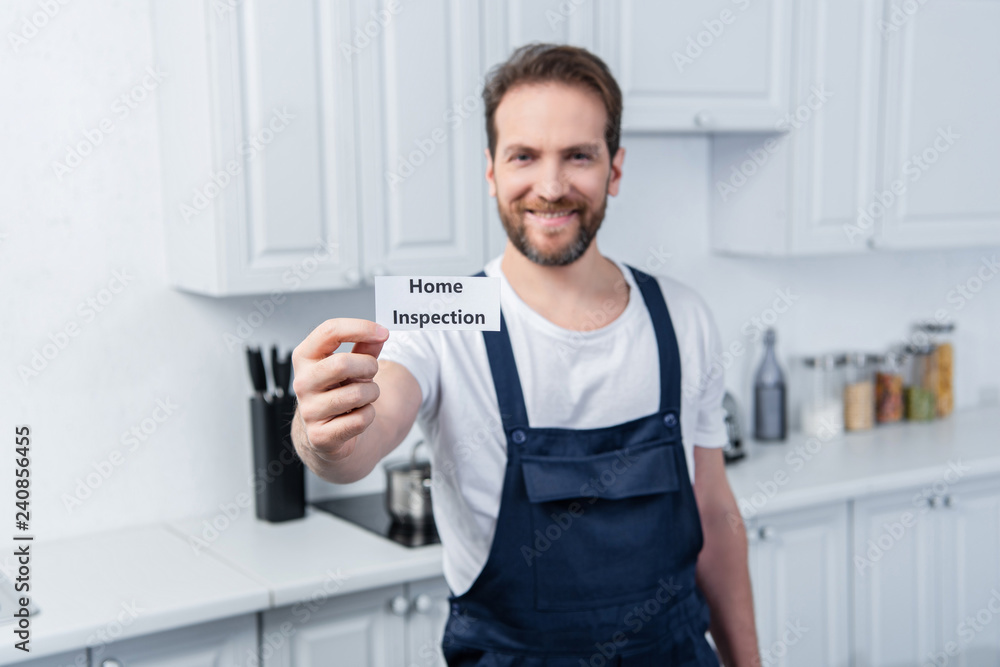 selective focus of happy male handyman in working overall showing card with lettering home inspection