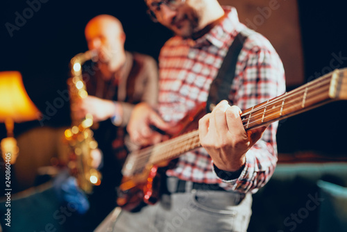 Close up of talented bass guitarist playing his instrumet. In background saxophonist playing sax. Home studioo interior.