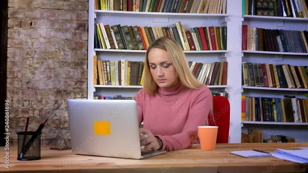 Portrait of concentrated caucasian young girl with piercing working attentively with laptop on book shelves background. Portrait of concentrated caucasian young girl with piercing working attentively with laptop on book shelves background.