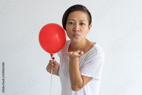 Asian woman holding red balloon and sending air kiss. Serious lady looking at camera and wearing t-shirt. Party and flirting concept. Isolated front view on white background. © Mangostar