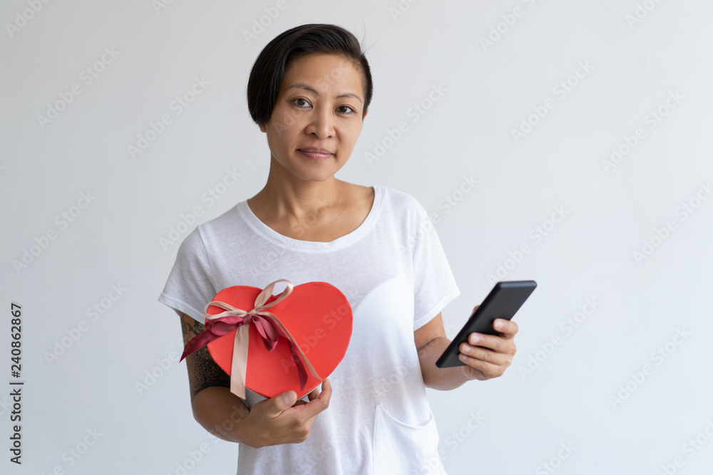 Content Asian woman holding heart shaped gift box and smartphone. Lady browsing on digital device. Valentines Day and communication concept. Isolated front view on white background. Content Asian woman holding heart shaped gift box and smartphone. Lady browsing on digital device. Valentines Day and communication concept. Isolated front view on white background.