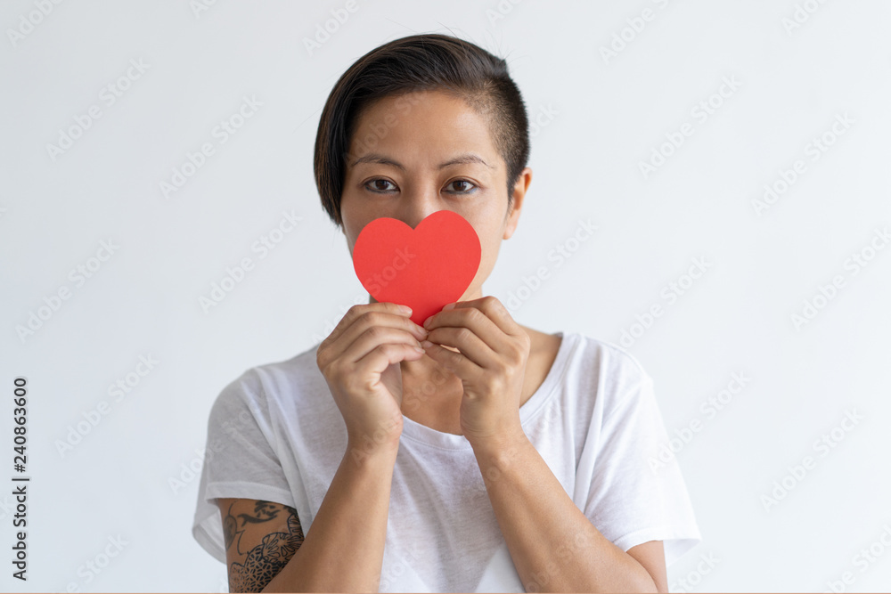 Playful Asian woman holding paper heart in front of her mouth. Tattooed lady wearing t-shirt and looking at camera. Saint Valentines Day concept. Isolated front view on white background. Playful Asian woman holding paper heart in front of her mouth. Tattooed lady wearing t-shirt and looking at camera. Saint Valentines Day concept. Isolated front view on white background.