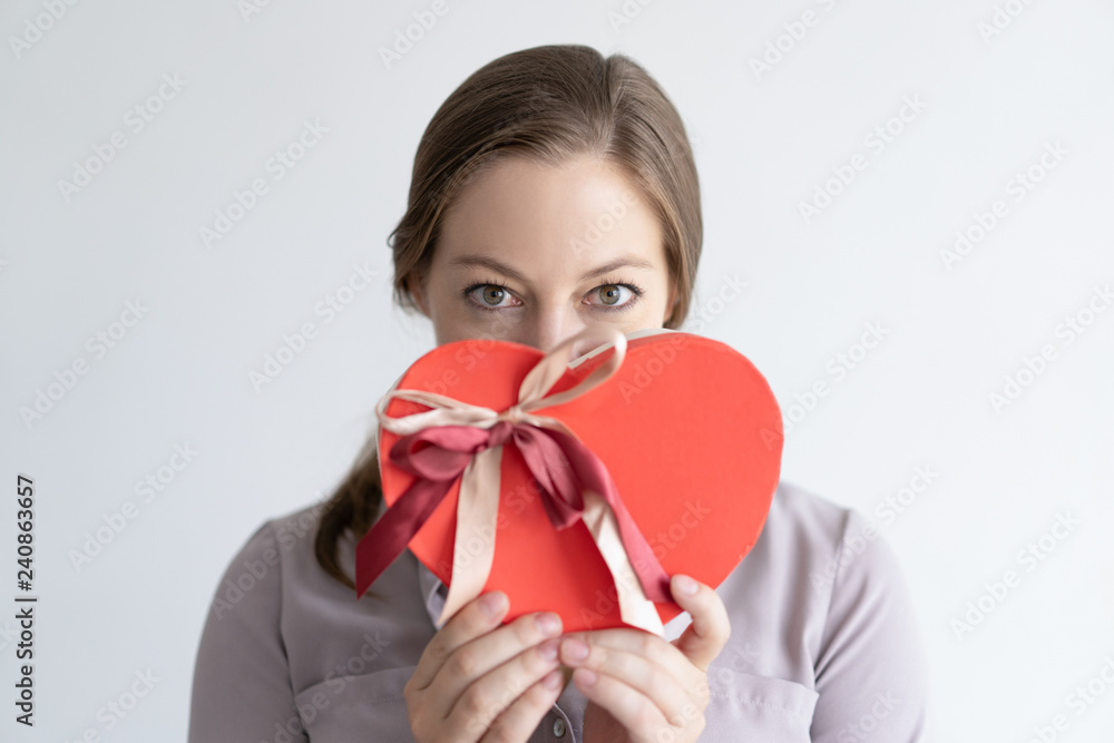 Playful lady holding heart shaped gift box in front of mouth. Woman wearing casual shirt and looking at camera. Saint Valentines Day gift concept. Isolated front view on white background. Playful lady holding heart shaped gift box in front of mouth. Woman wearing casual shirt and looking at camera. Saint Valentines Day gift concept. Isolated front view on white background.