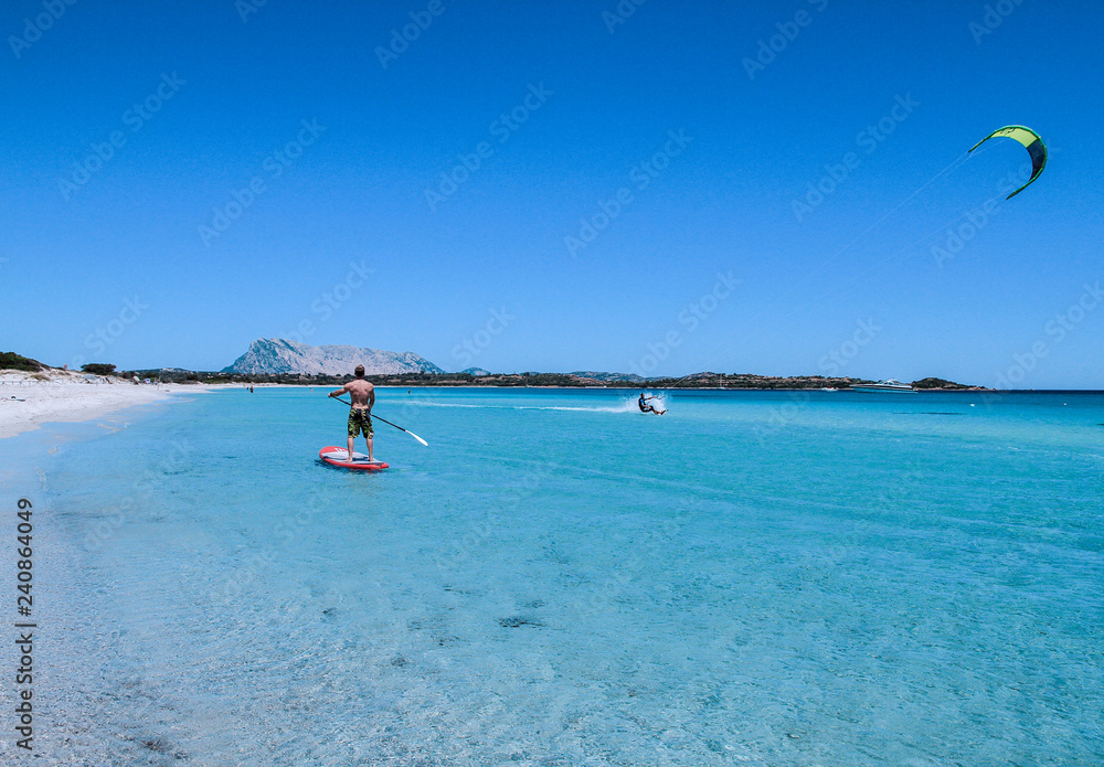 A young athlete paddle on stand up paddle on the crystalline sea of Sardinia, while a kitesurfer slides on the water beside him