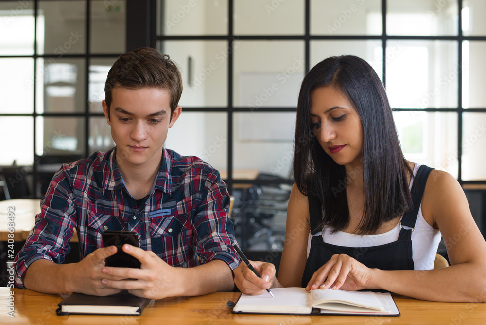 Portrait of concentrated student girl and boy sitting at desk. Young Caucasian man using mobile phone and woman writing in notebook. Education concept Portrait of concentrated student girl and boy sitting at desk. Young Caucasian man using mobile phone and woman writing in notebook. Education concept