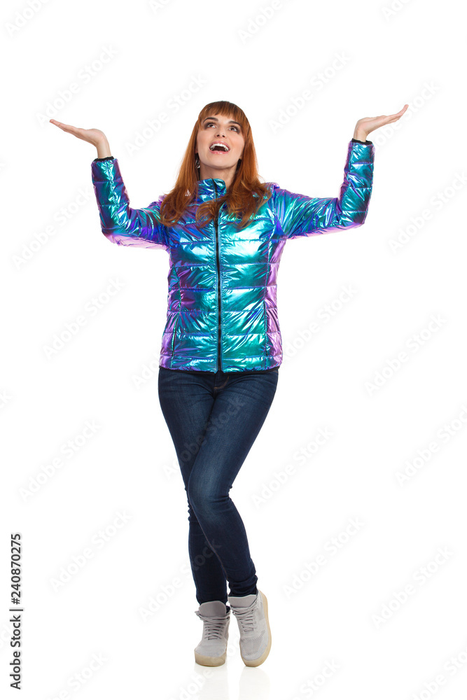 Young Woman In Vibrant Down Jacket Is Holding Hands Raised, Showing Something And Looking Up Young Woman In Vibrant Down Jacket Is Holding Hands Raised, Showing Something And Looking Up
