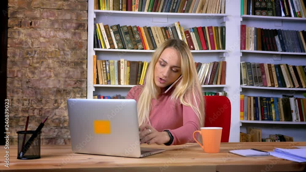 Portrait of young caucasian girl with piercing attentively working with laptop and talking on cellphone on book shelves background. Portrait of young caucasian girl with piercing attentively working with laptop and talking on cellphone on book shelves background.