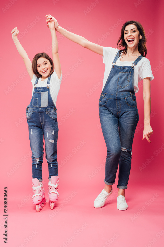 Fototapeta premium happy daughter in rollerblades cheering and holding hands with mother on pink background
