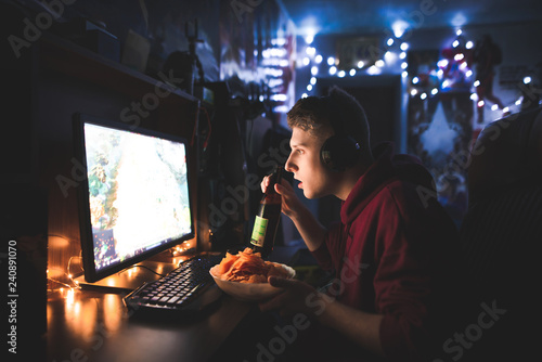 Young man uses a computer at home in a cozy room, eats chips, drinks beer and looks focused on the screen. Gamer port plays home games and eats snacks. Fastfood dinner at the computer
