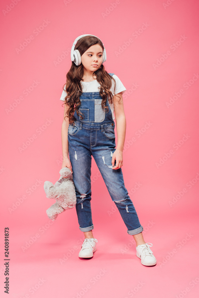 full length of sad kid standing in headphones and holding teddy bear on pink background full length of sad kid standing in headphones and holding teddy bear on pink background