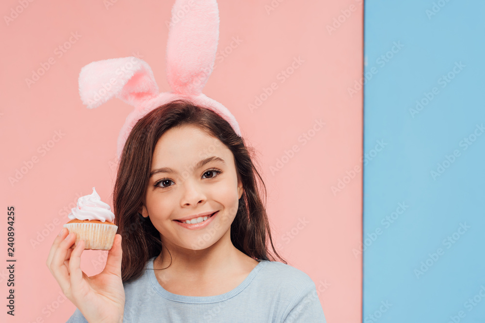 adorable child in bunny ears holding cupcake and looking at camera on blue and pink background adorable child in bunny ears holding cupcake and looking at camera on blue and pink background