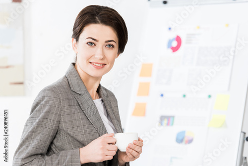 cheerful beautiful businesswoman in grey suit holding cup of coffee in office