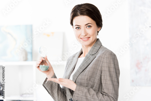 smiling attractive businesswoman in grey suit showing hourglass and looking at camera in office